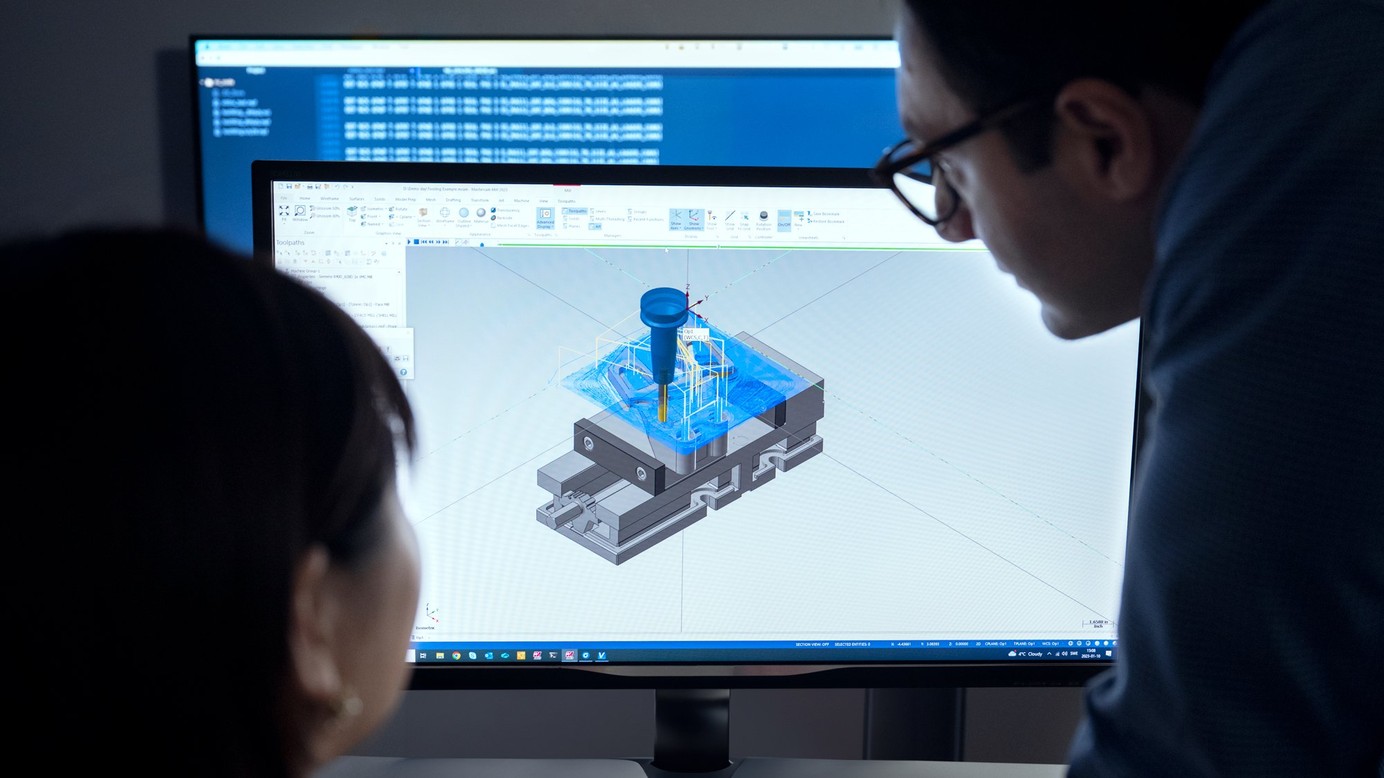 A woman and a man looking at a computer screen with an image of a toolpath for a milling operation which is generated using CAM software