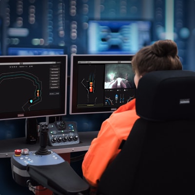 A woman sitting at a desk in a control room looking into two monitors showing different views of a mine