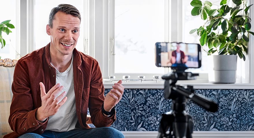 A man looking in to a mobile phone on a stand while the camera is filming him